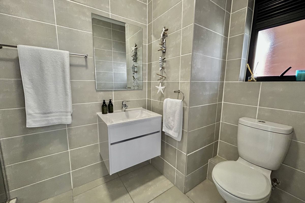Modern bathroom featuring neutral grey wall and floor tiling, a wall-mounted vanity with basin, and a dual-flush toilet.