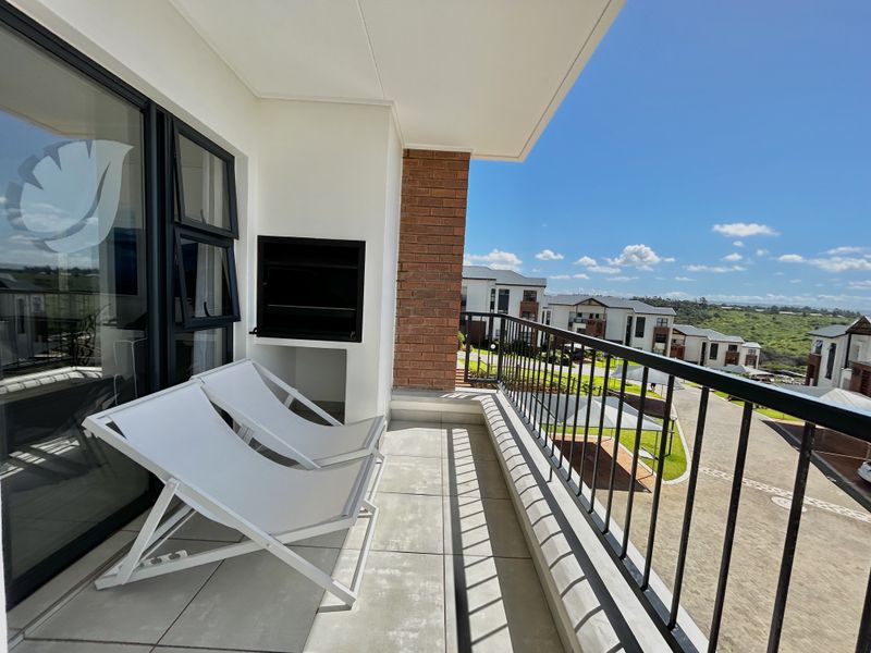 A modern covered balcony featuring a built-in braai, tiled flooring, and a metal balustrade, offering pleasant views of the surrounding estate.