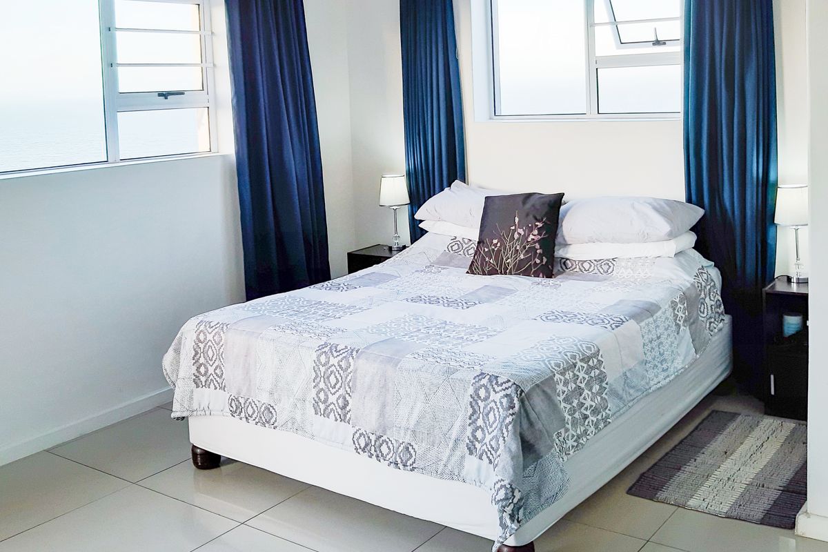 A bright bedroom featuring large windows with ocean views, neutral tiled flooring, and clean white walls.