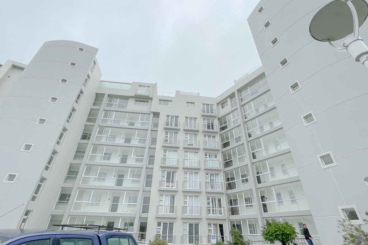 Low-angle exterior view of a modern multi-story apartment building featuring white facade, numerous balconies, and large glass windows.