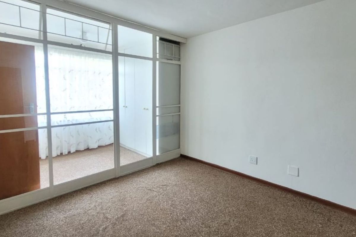A vacant bedroom featuring dated carpeting, built-in cupboards, and a large glass partition wall leading to an enclosed balcony area.