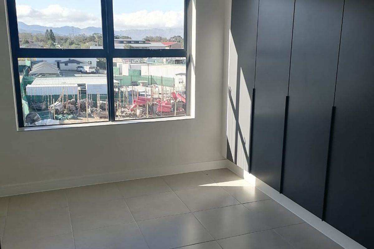 A bright, modern bedroom featuring sleek dark built-in cupboards, neutral floor tiles, and a large window with views of the surrounding area.