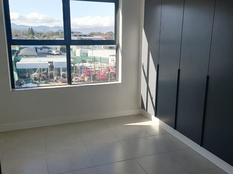 A bright, modern bedroom featuring sleek dark built-in cupboards, neutral floor tiles, and a large window with views of the surrounding area.