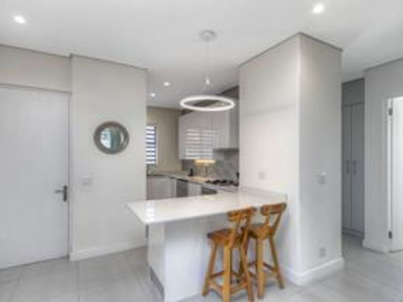 Modern open-plan kitchen featuring sleek white cabinetry, stone countertops, a breakfast bar, and integrated lighting, seamlessly flowing into the living area.