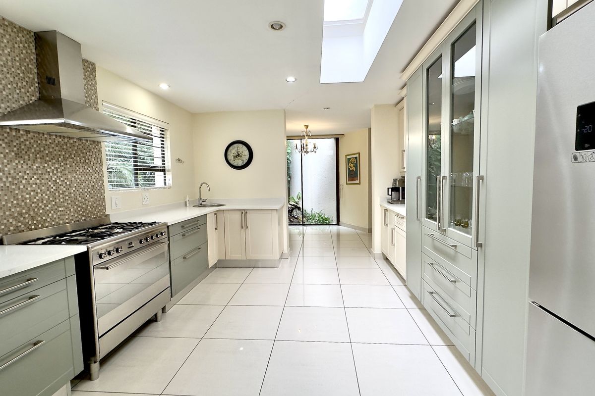 Modern kitchen featuring light-toned cabinetry, stone countertops, a stainless steel gas range with extractor hood, tiled flooring, and a bright skylight.