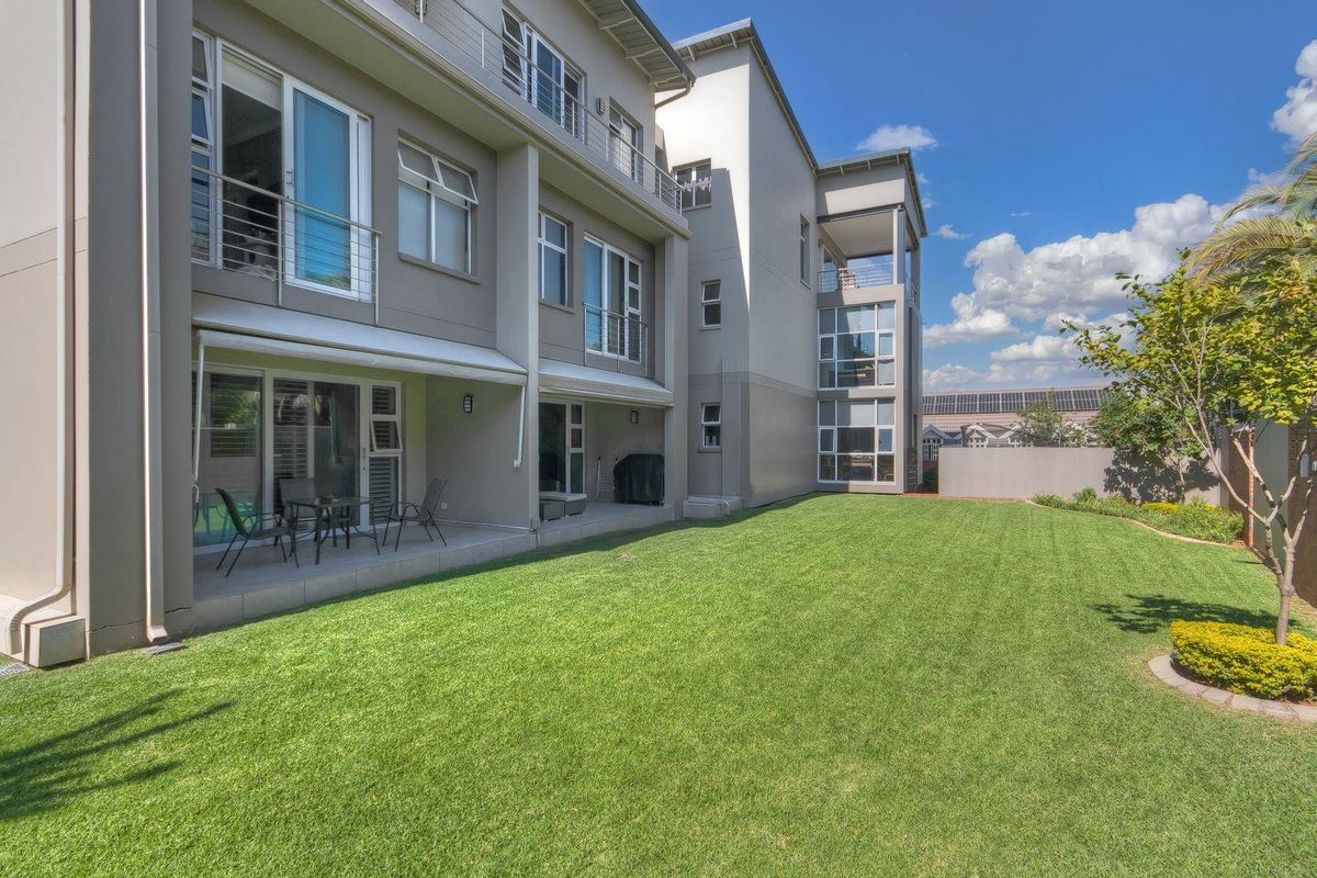 A well-maintained rear exterior view of the apartment building featuring a paved patio area, sliding glass doors, and a spacious private lawn.