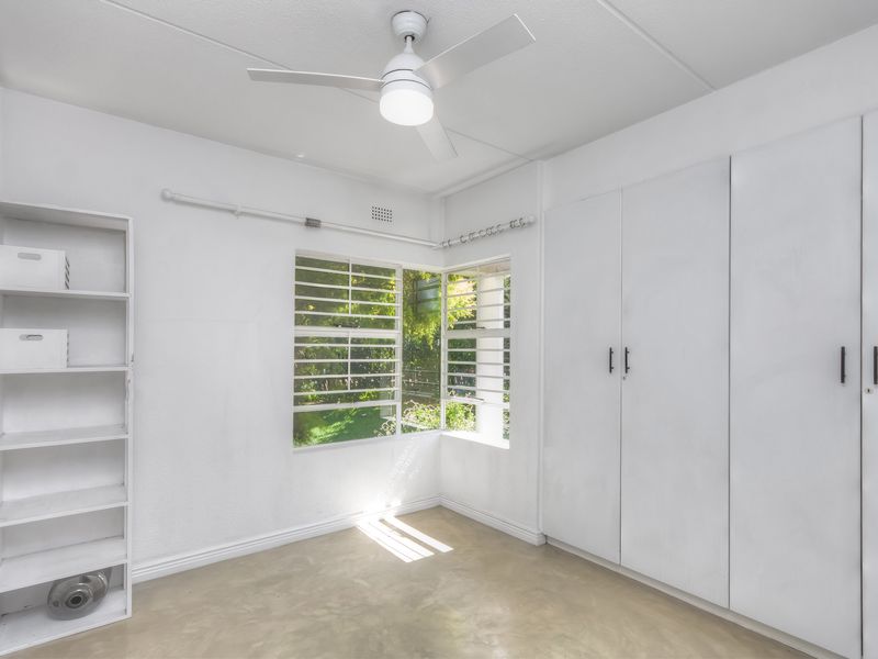 A bright, empty bedroom featuring built-in cupboards, a ceiling fan, and louvered windows overlooking a garden area.
