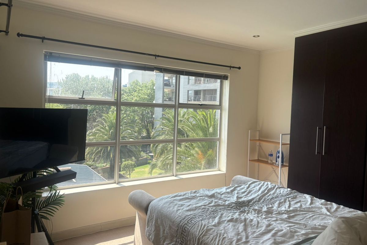 A well-lit bedroom featuring a large window with garden views, built-in dark wood wardrobe, recessed ceiling lighting, and neutral carpet flooring.