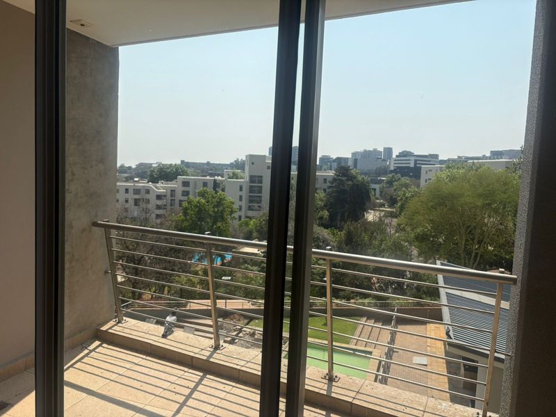A private balcony featuring a stainless steel balustrade and tiled flooring, offering an elevated view of the surrounding apartment complex and greenery.