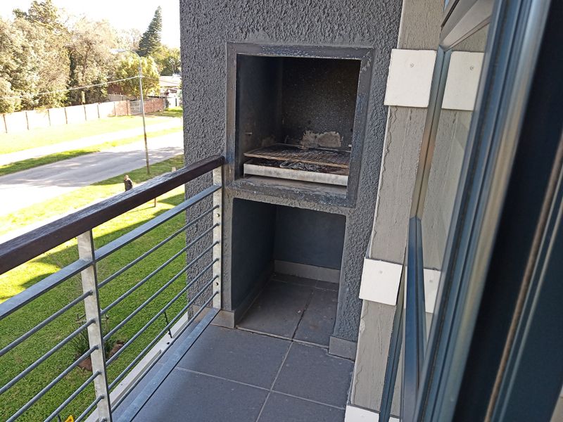 A balcony featuring a built-in braai, tiled flooring, and metal railings, overlooking a street-side green space.
