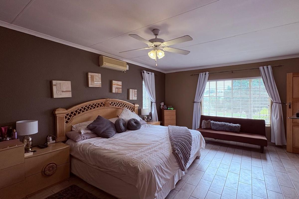 Spacious bedroom featuring tiled flooring, a ceiling fan, and a wall-mounted air conditioning unit with large windows and venetian blinds.