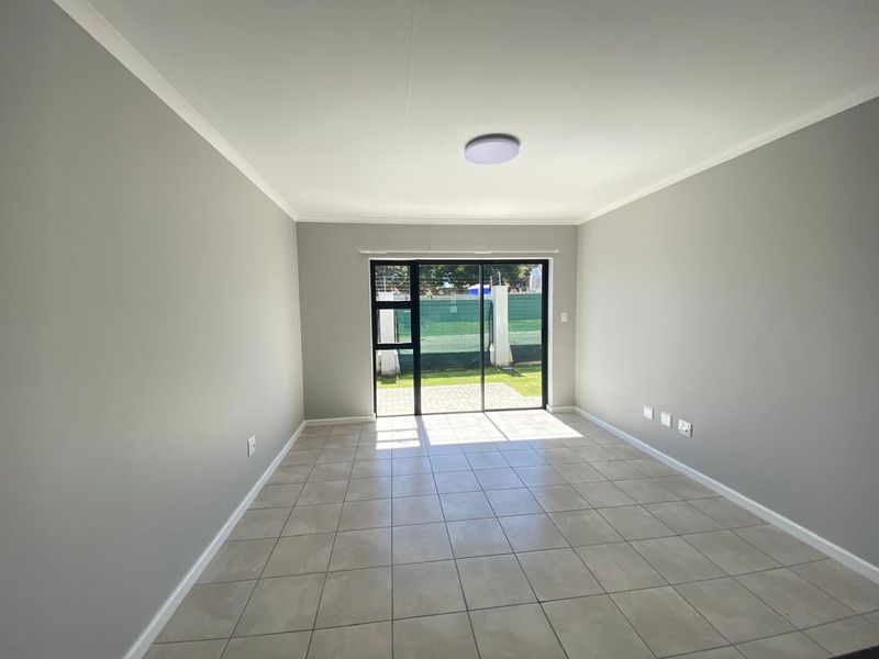 A bright, empty living room featuring neutral grey walls, tiled flooring, and a sliding glass door providing access to the outdoor area.