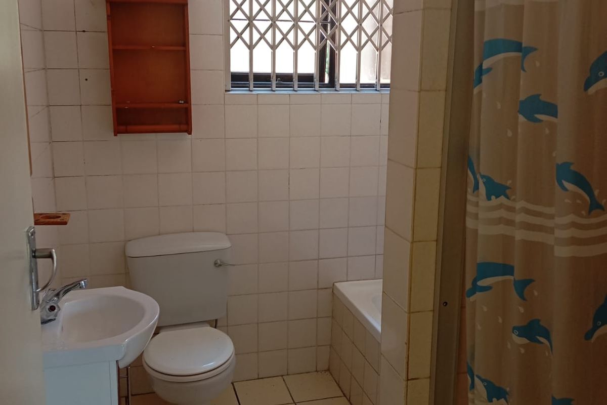 A functional bathroom featuring a standard bathtub, pedestal basin, and toilet, finished with white wall and floor tiles and a security-barred window.