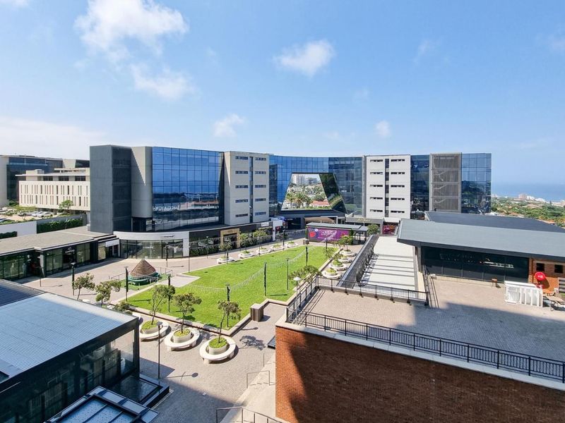 An elevated aerial view of a modern commercial complex featuring a central landscaped courtyard, glass-fronted buildings, and paved outdoor areas.