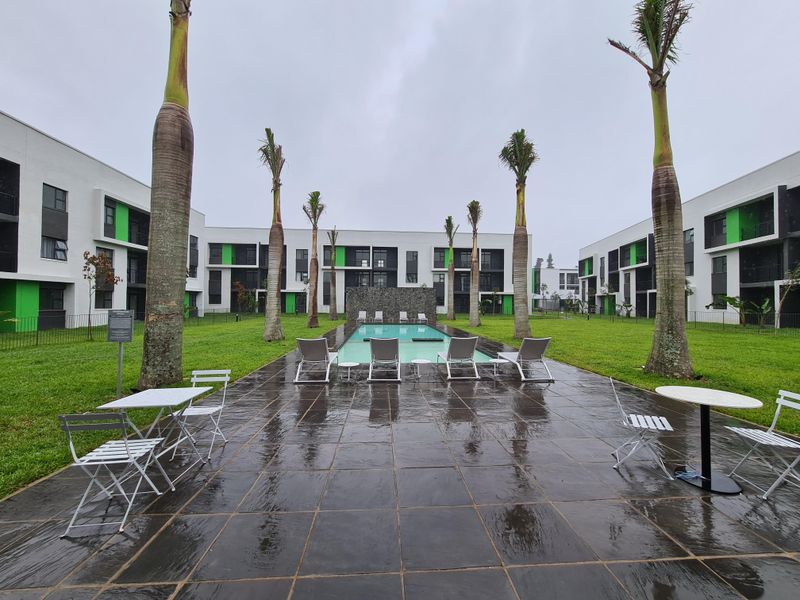 Communal swimming pool area featuring a stone feature wall, paved patio, and landscaped lawn surrounded by modern apartment buildings.