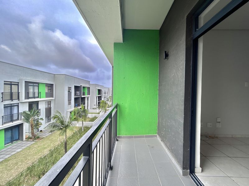 A modern apartment balcony featuring tiled flooring, a metal balustrade, and a vibrant green accent wall, overlooking the complex grounds.