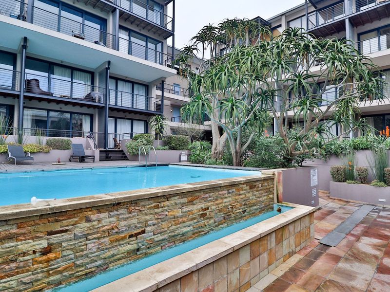 Communal swimming pool area featuring stone-clad water feature, timber decking, and landscaped gardens set within a modern apartment complex.
