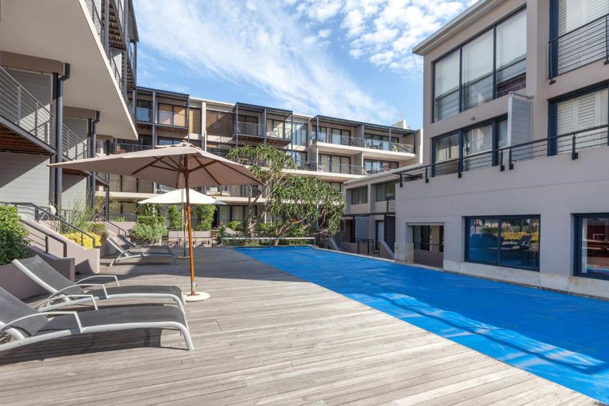 A well-maintained communal swimming pool area featuring timber decking, surrounded by modern apartment blocks with balconies and large glass sliding doors.