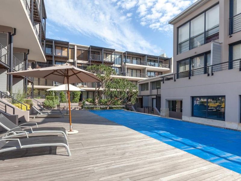 A well-maintained communal swimming pool area featuring timber decking, surrounded by modern apartment blocks with balconies and large glass sliding doors.