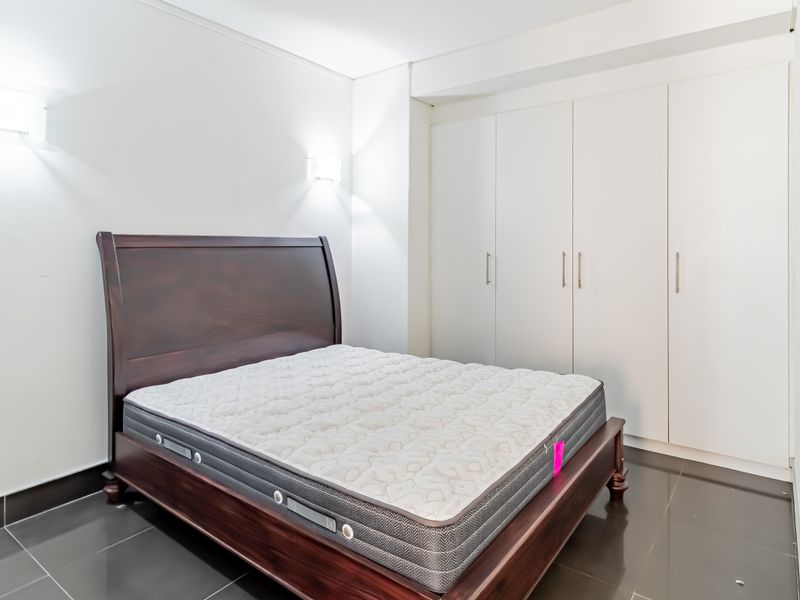 A bright bedroom featuring dark tiled flooring, built-in white cupboards, and modern wall-mounted lighting.