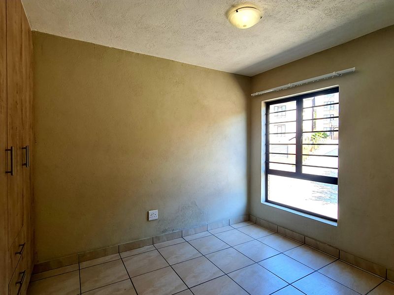 A vacant bedroom featuring tiled flooring, built-in wooden cupboards, and a window fitted with security bars.