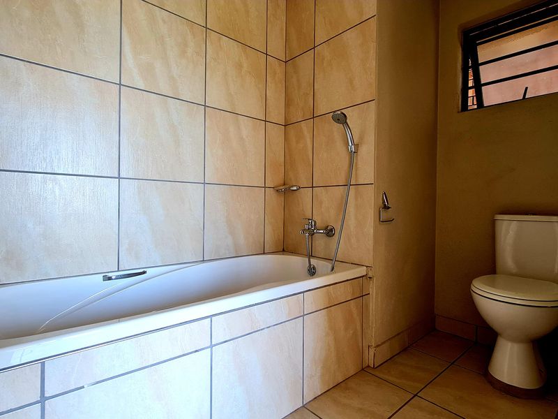 A functional bathroom featuring a built-in bathtub with shower mixer, a standard toilet, and neutral wall and floor tiling.