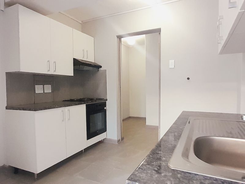 A functional, empty kitchen featuring white cabinetry, dark laminate countertops, a stainless steel sink, and built-in oven with gas hob and extractor fan.