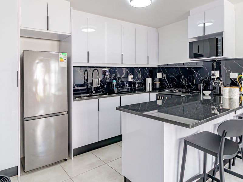 Modern kitchen featuring high-gloss white cabinetry, black marble-look splashback, granite-look countertops, a functional kitchen island, and integrated appliances.