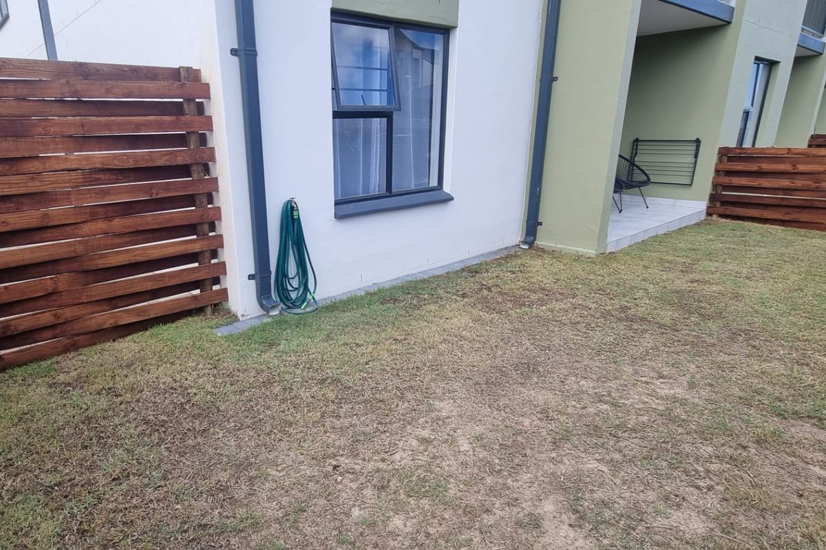 A private rear garden area featuring timber slat fencing, a small tiled patio, and established lawn space adjacent to the apartment building.