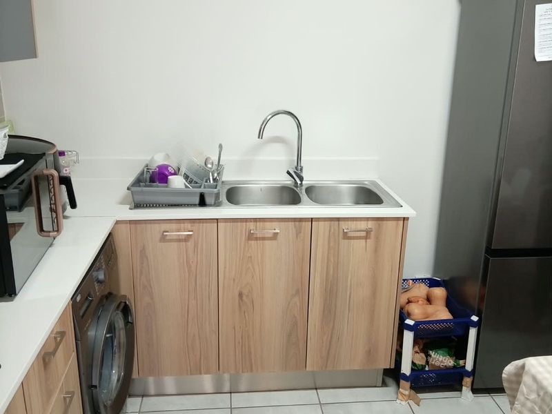 A functional kitchen area featuring light wood-finish cabinetry, a double bowl sink with mixer tap, tiled flooring, and integrated laundry and cooking appliances.