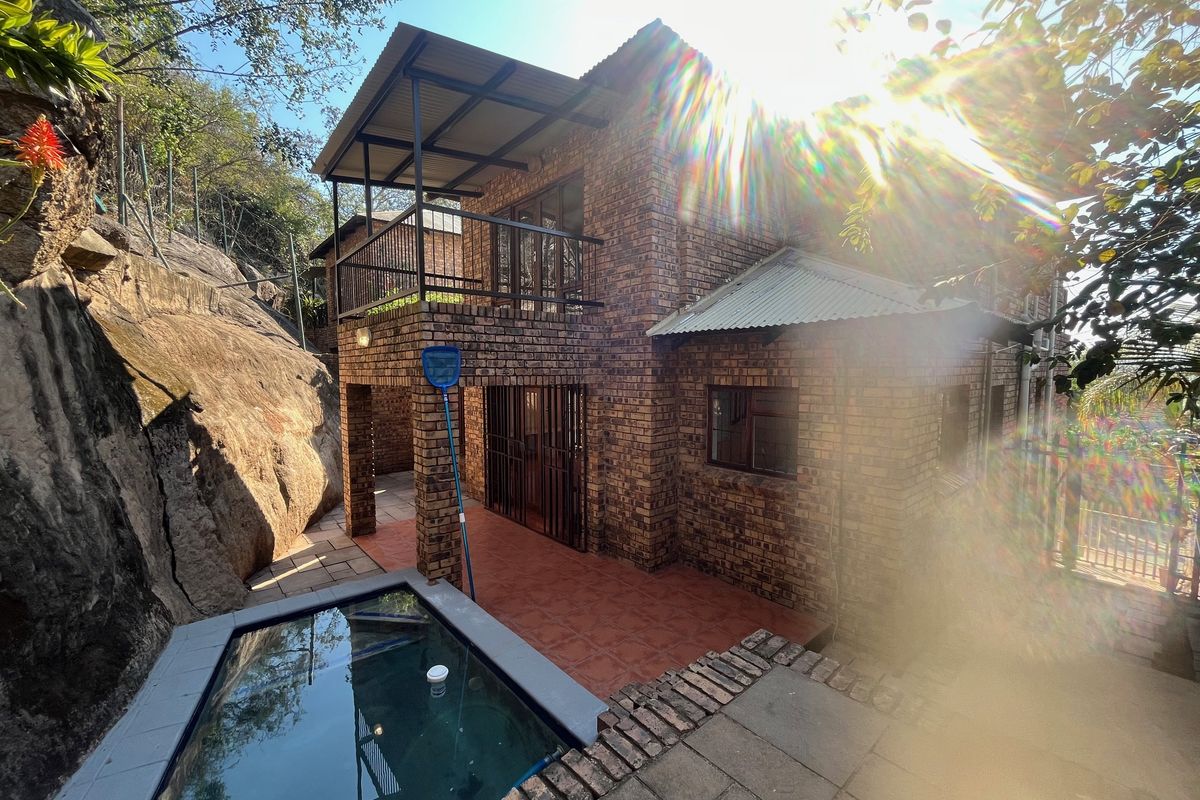 Rear view of a face brick house featuring a balcony, tiled patio area, and a small swimming pool set against a natural rock formation.