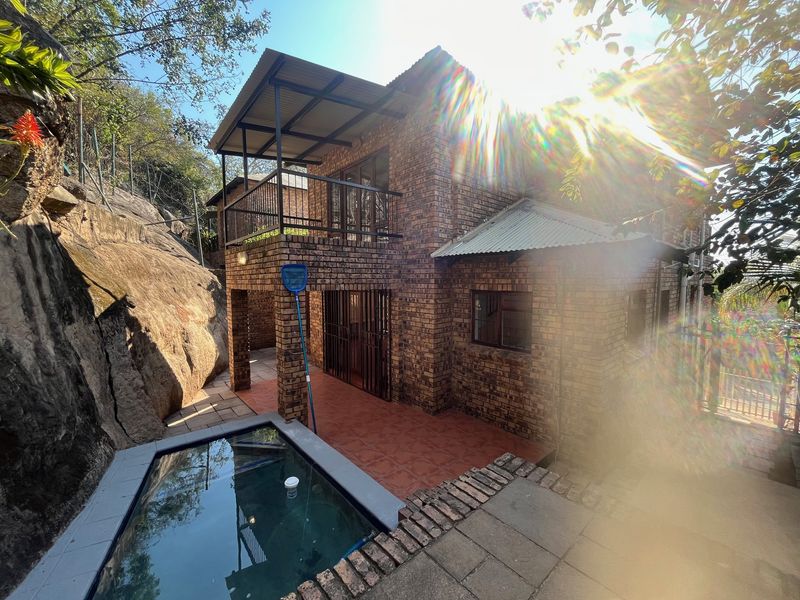 Rear view of a face brick house featuring a balcony, tiled patio area, and a small swimming pool set against a natural rock formation.