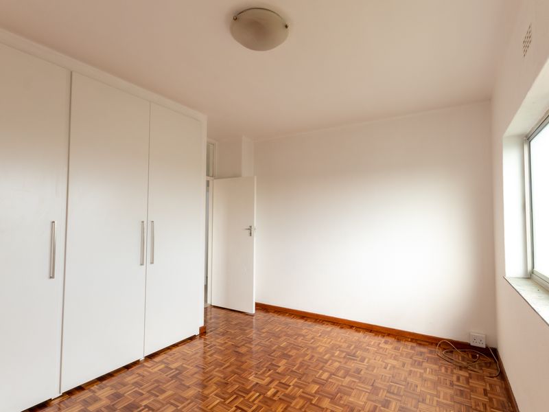 A bright, empty bedroom featuring classic parquet flooring, white built-in cupboards, and a large window providing natural light.