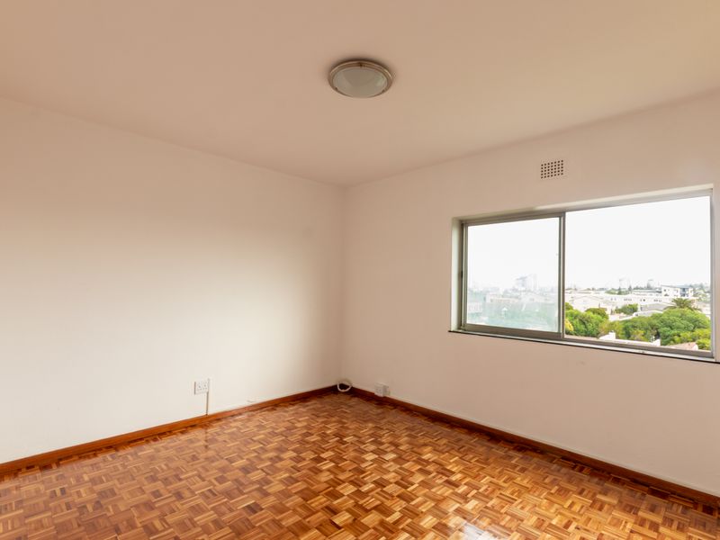 A bright, empty bedroom featuring well-maintained parquet flooring, a large sliding window with an urban view, and neutral wall finishes.