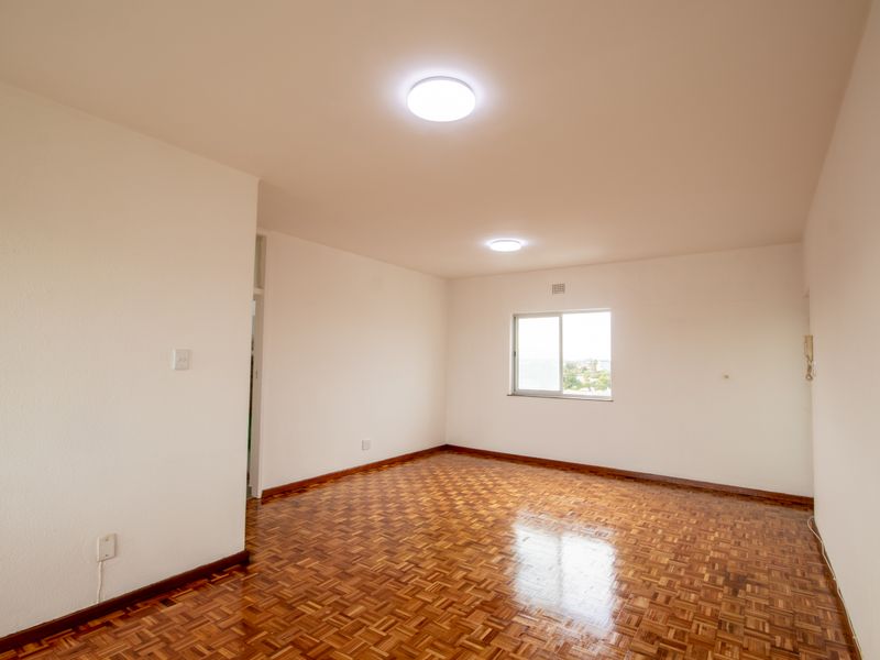 A bright, empty living room featuring polished parquet flooring, neutral walls, and a sliding window providing natural light.