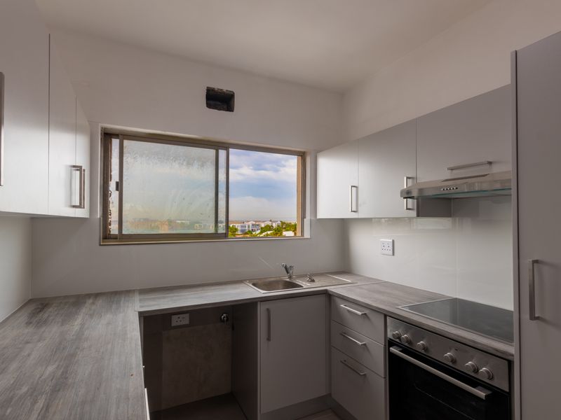 A compact, modern kitchen featuring white cabinetry, grey laminate countertops, integrated oven and hob, and a window with an exterior view.