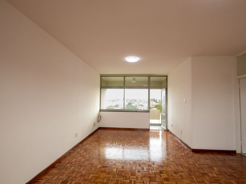 An empty living room featuring polished parquet flooring, neutral walls, and a sliding glass door leading to a balcony with neighborhood views.