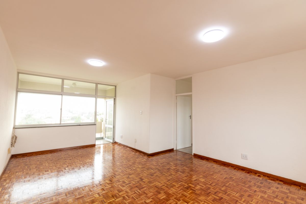 A bright, empty living room featuring polished parquet flooring, large aluminium-framed windows, and neutral walls, offering a clean canvas for interior design.