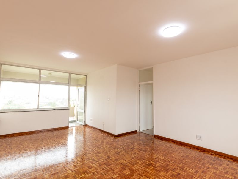 A bright, empty living room featuring polished parquet flooring, large aluminium-framed windows, and neutral walls, offering a clean canvas for interior design.