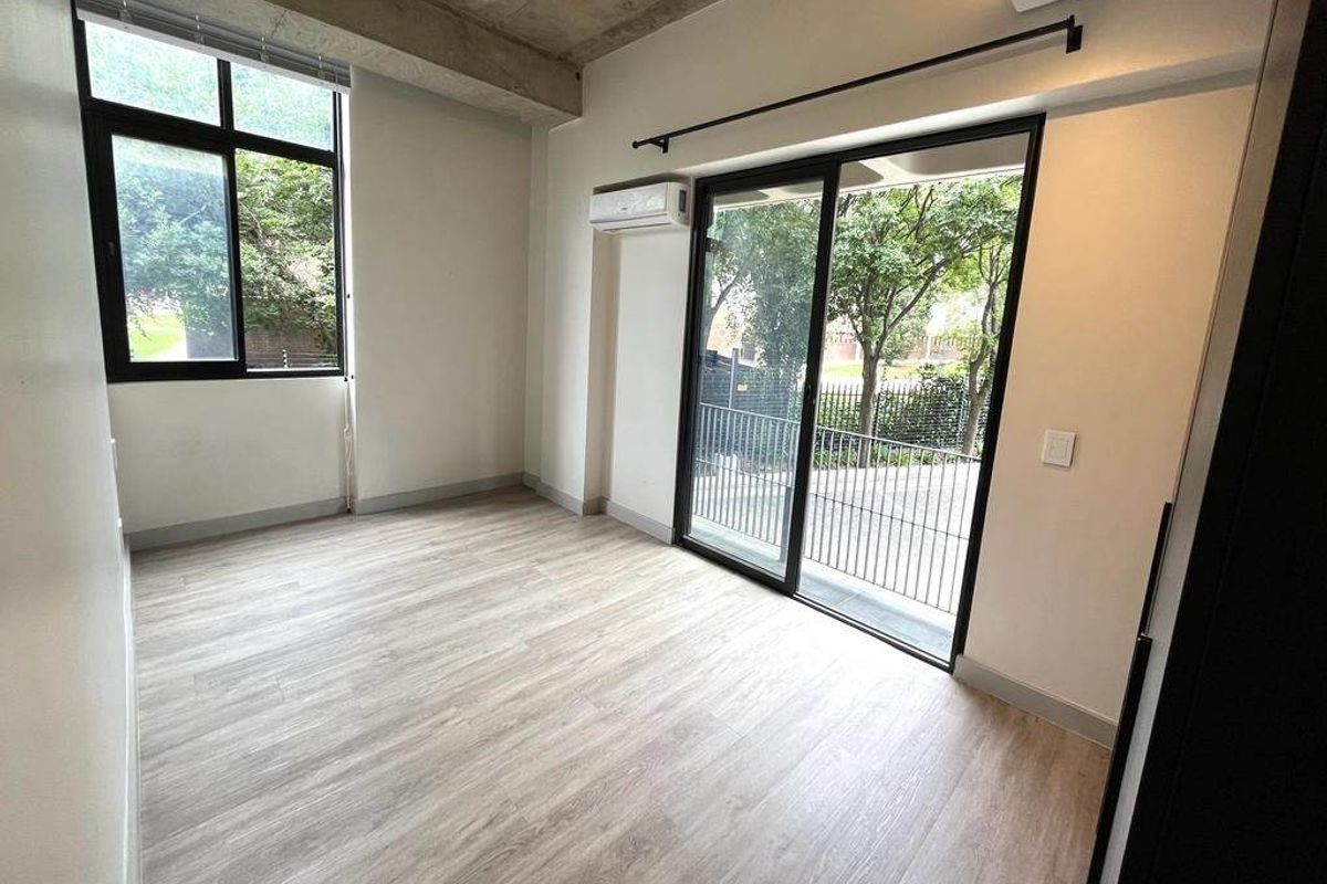 A bright, modern bedroom featuring light wood-look flooring, exposed concrete ceiling beams, a wall-mounted air conditioning unit, and sliding glass doors leading to a balcony.