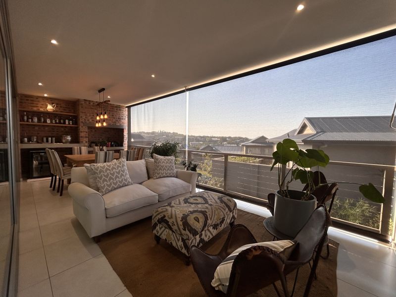 Spacious covered balcony featuring a built-in braai, exposed brick accents, modern recessed lighting, and a glass balustrade with expansive neighborhood views.