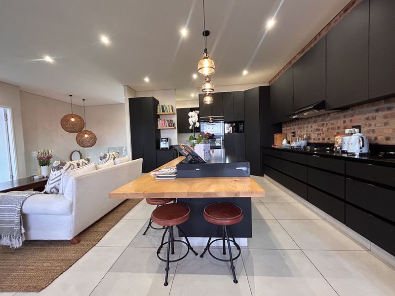 Modern open-plan kitchen and living area featuring sleek black cabinetry, a large central island with wooden breakfast bar, and an exposed brick feature wall.