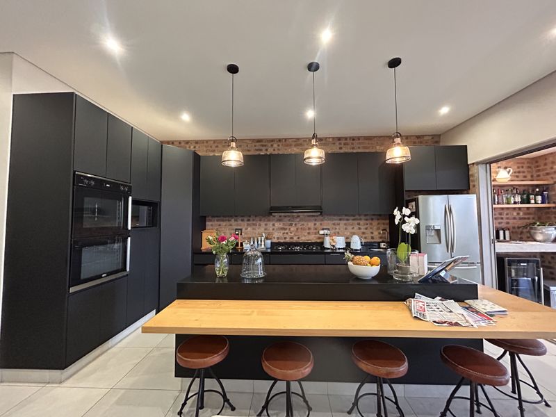 Modern kitchen featuring sleek dark cabinetry, an exposed brick backsplash, a central island with a wooden breakfast bar, and high-end built-in appliances.