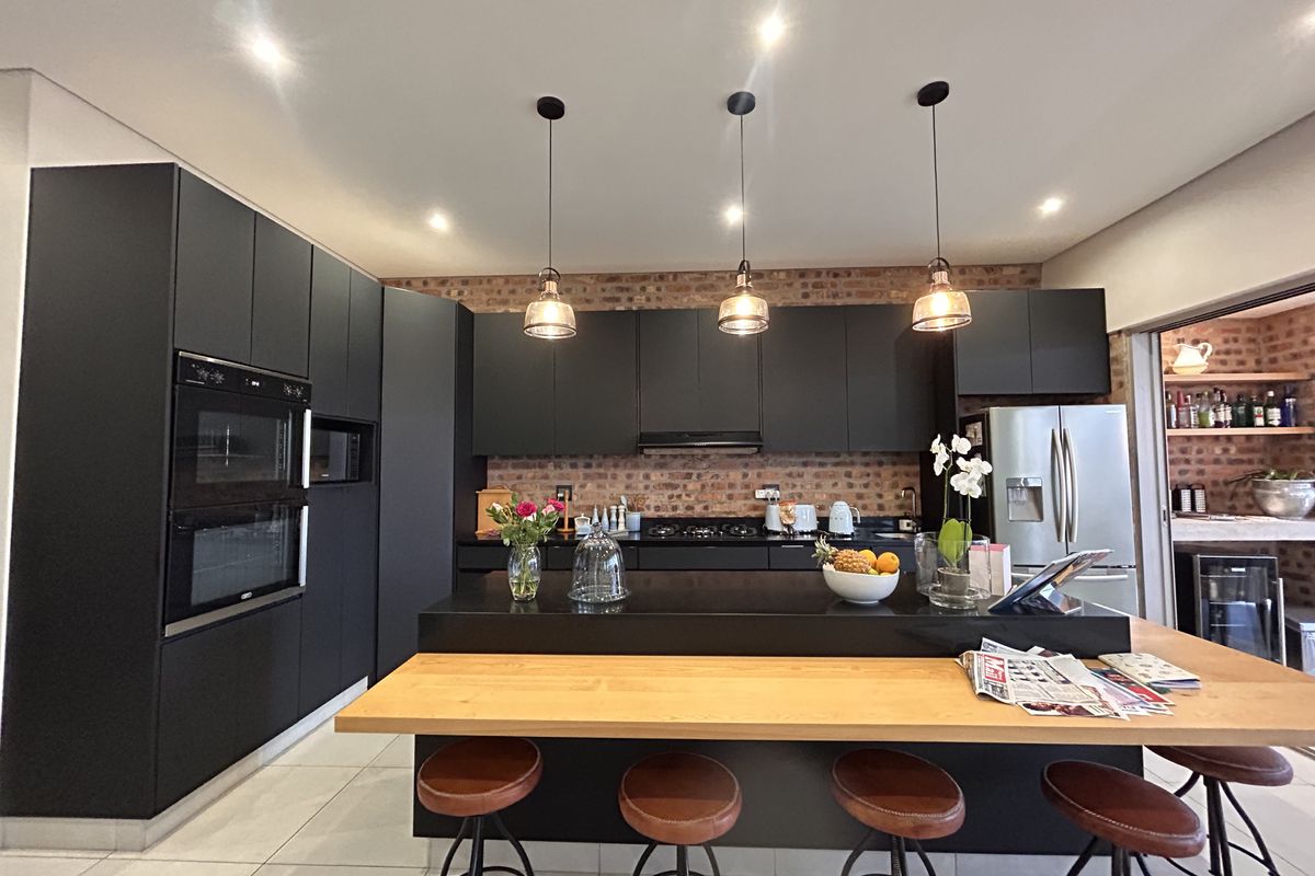 Modern kitchen featuring sleek dark cabinetry, an exposed brick backsplash, a central island with a wooden breakfast bar, and high-end built-in appliances.