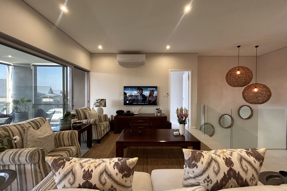 A well-lit living room featuring recessed downlights, a split-system air conditioner, and a modern glass balustrade overlooking the staircase.