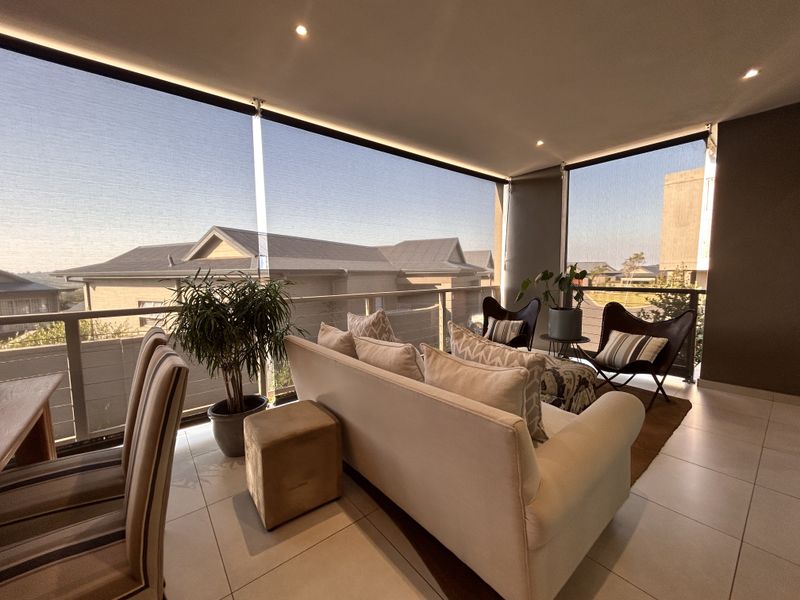 A spacious covered balcony featuring tiled flooring, recessed lighting, and retractable roller blinds, offering a comfortable outdoor living area with neighborhood views.