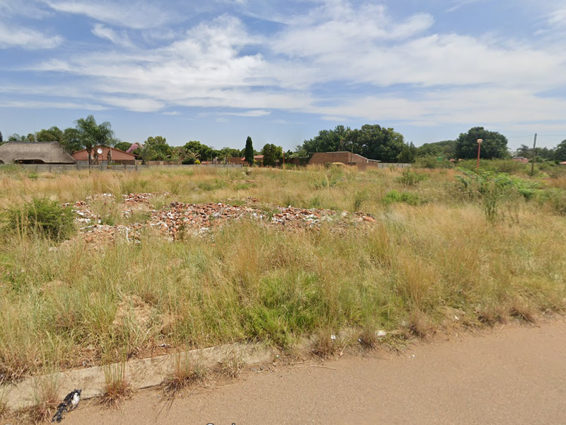 A vacant residential plot featuring overgrown grass and a small pile of rubble, situated along a paved road.