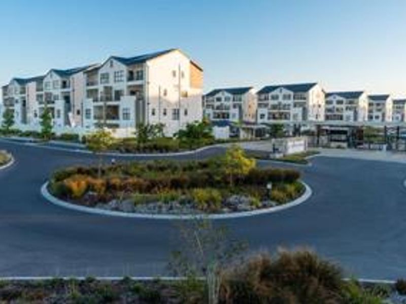 A wide-angle view of the apartment complex entrance, featuring a landscaped roundabout, paved driveway, and modern multi-story residential buildings.