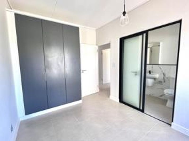 A modern, empty bedroom featuring sleek dark built-in cupboards, neutral tiled flooring, and a sliding glass door leading to an en-suite bathroom.