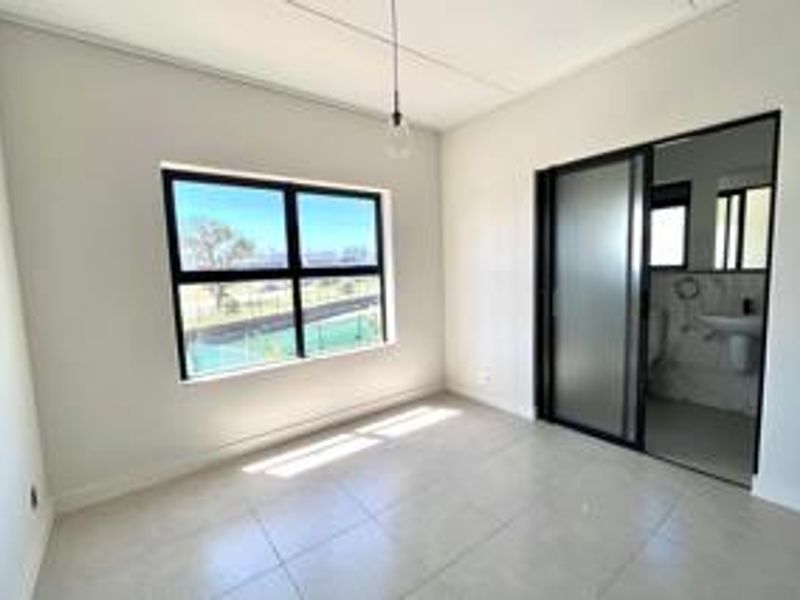 A bright, modern bedroom featuring neutral tiled flooring, large black-framed windows, and a sleek sliding door leading to an en-suite bathroom.